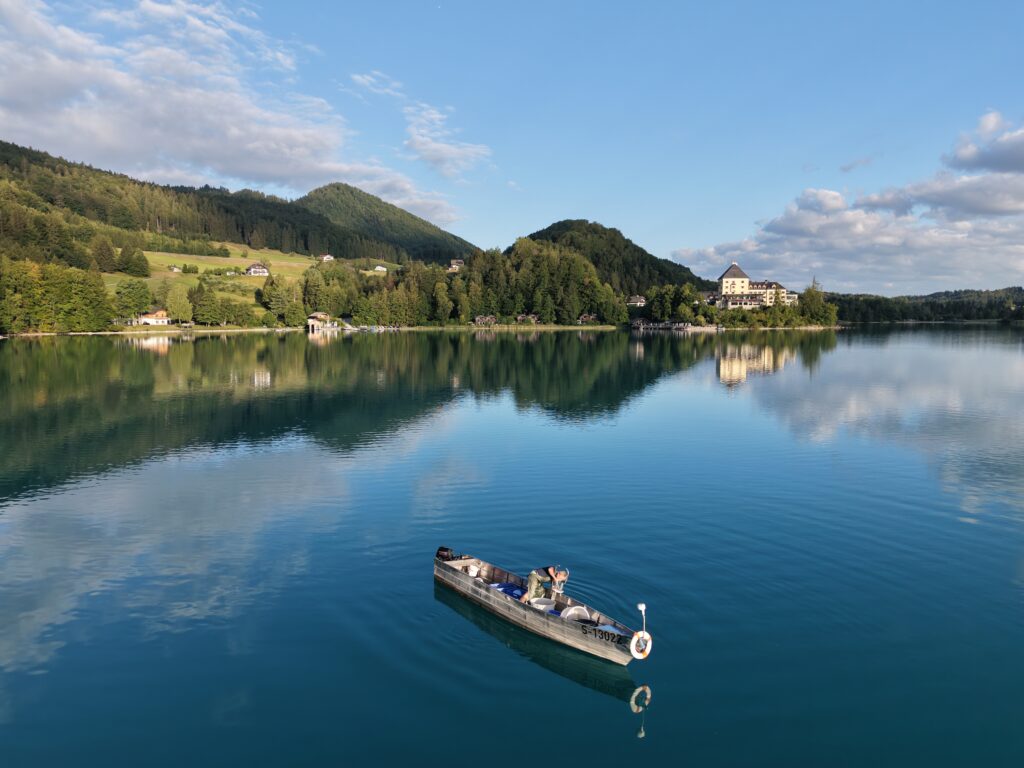 Fischer fischt am Fuschlsee im Hintergrund das Rosewood Schloss Fuschl und die Berge im Salzkammergut